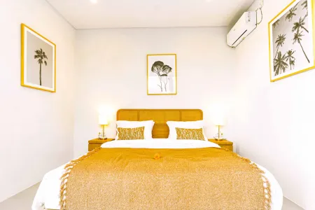 Cozy minimalist bedroom with king-sized tan-upholstered bed, gold fringe blanket, palm tree wall art, and bedside lamps in a tropical style.